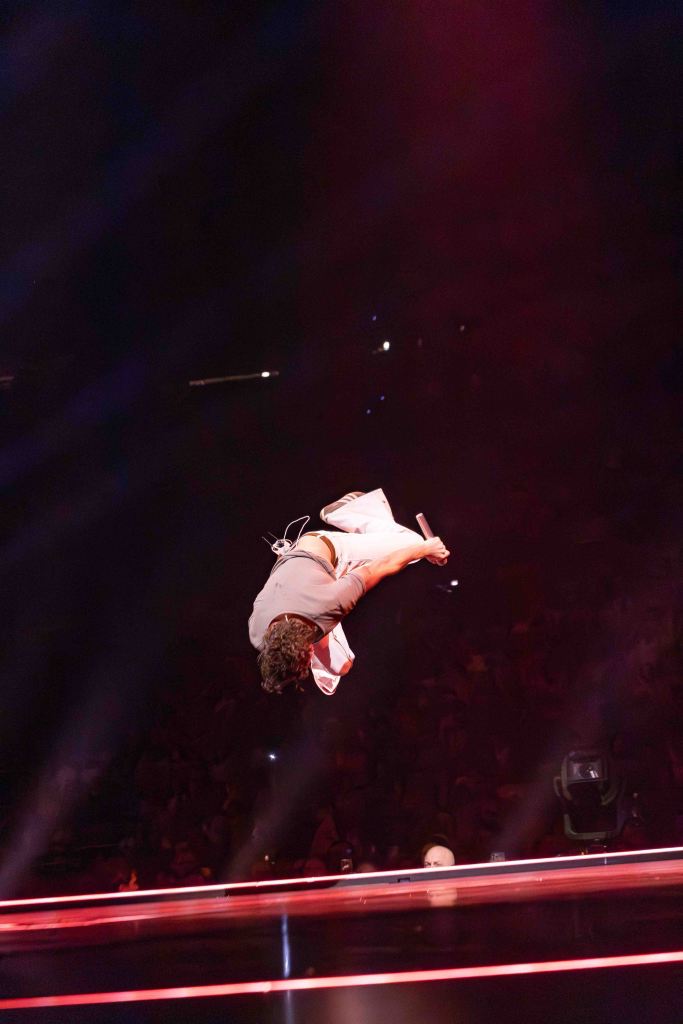 A performer executing a mid-air flip on stage, holding a microphone, with dramatic lighting and a blurred audience in the background.