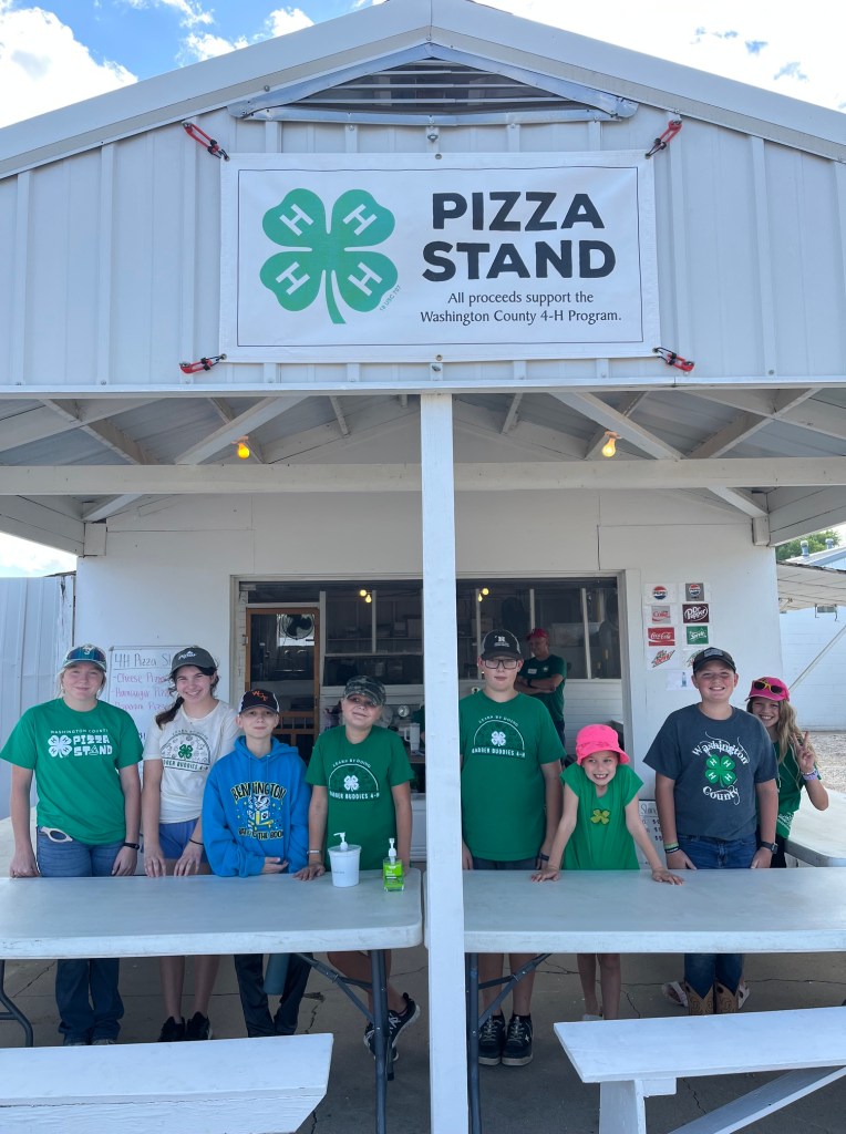 A group of children wearing green 4-H shirts standing in front of a pizza stand at an event, with a sign that reads 'Pizza Stand' above them.