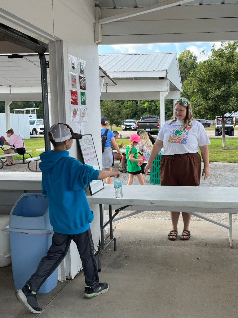 A child in a blue hoodie is handing a water bottle to a vendor at a food stand. The vendor, wearing a floral-themed shirt, is smiling while standing behind a table. In the background, other people are gathered around the stand in a park-like setting.