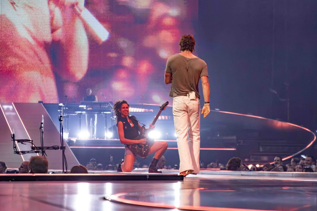 A female guitarist performing on stage while a male singer faces her, with a vibrant background and an engaged audience.