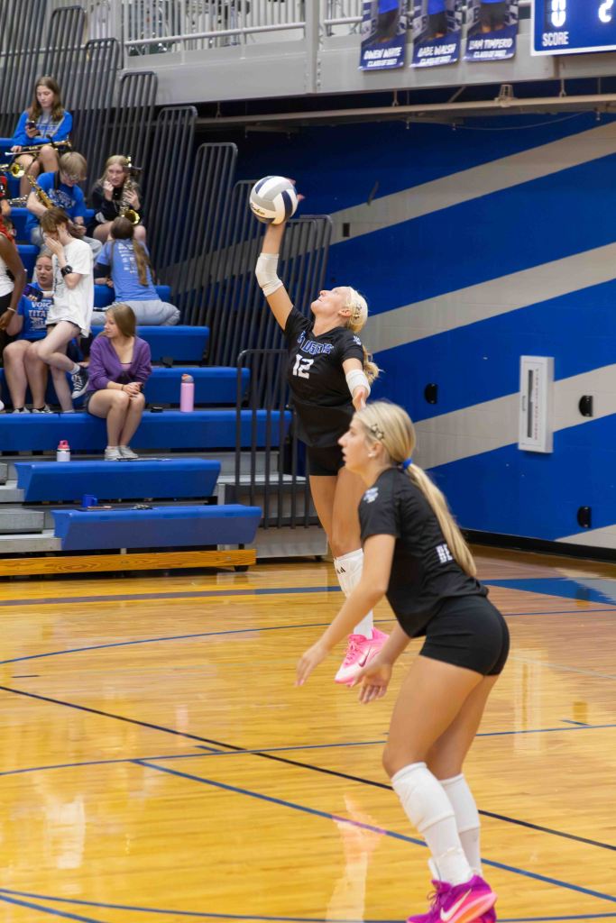 A volleyball player in a black uniform serving the ball during a match, with teammates and spectators in the background.