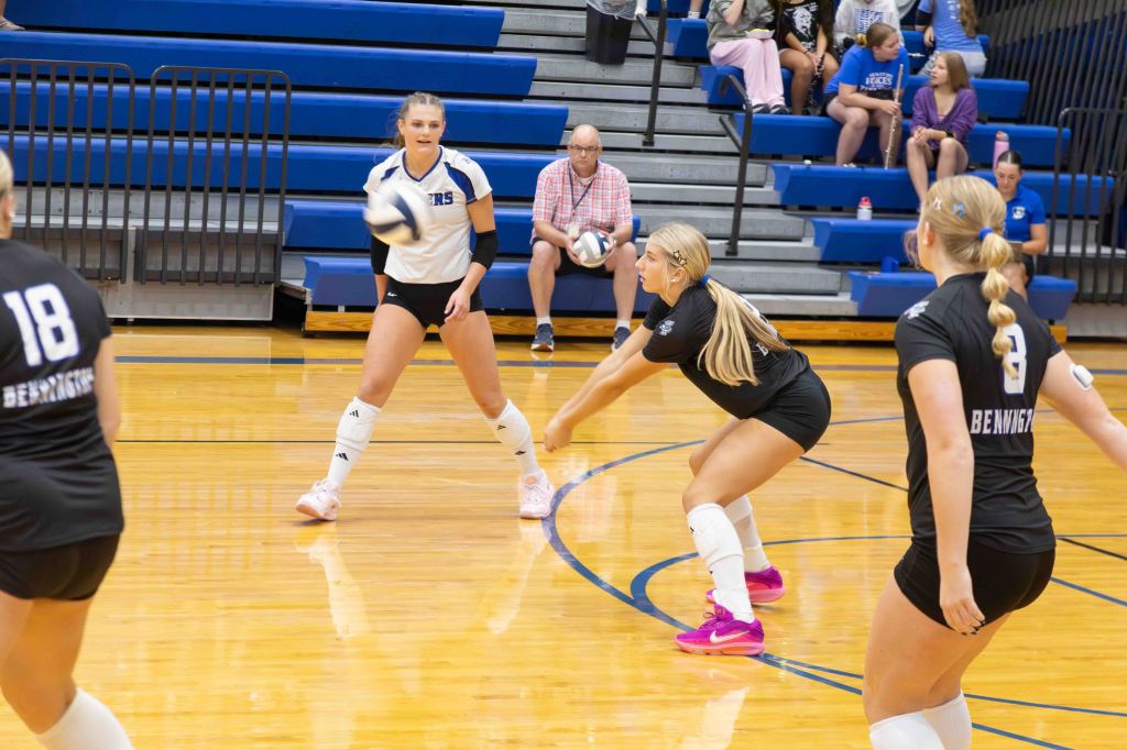 A volleyball game in progress with players on the court. One player, wearing a white jersey, is about to set the ball, while another player in a black jersey prepares to receive it. Spectators are visible in the background.