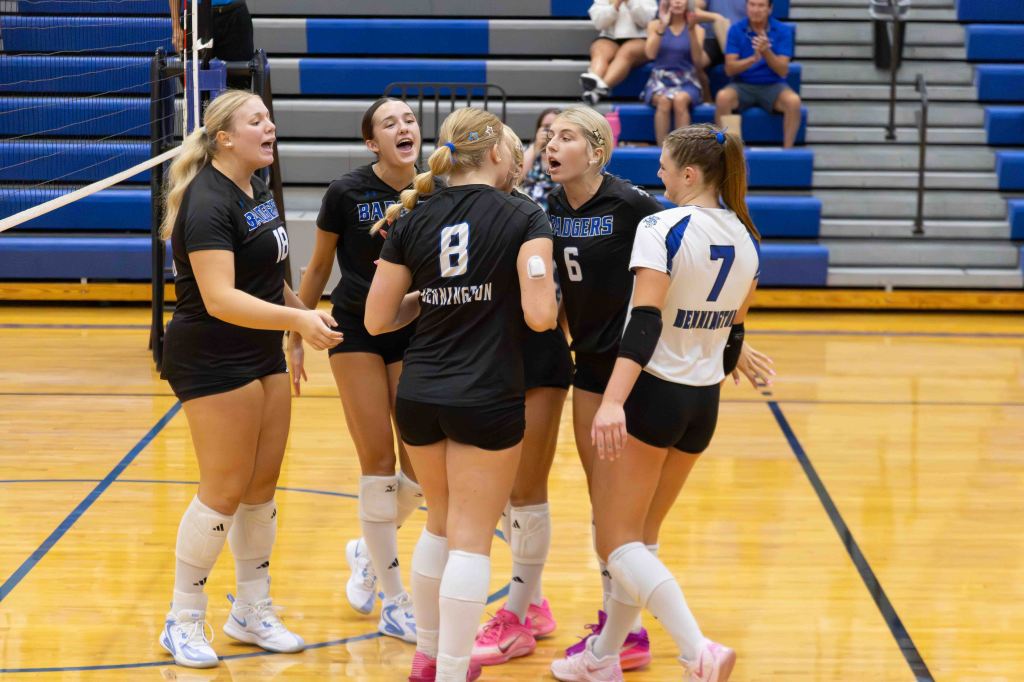 A group of five female volleyball players huddled together, celebrating and cheering on the court. They are wearing black and white uniforms, with visible team names. The background shows bleachers filled with spectators.