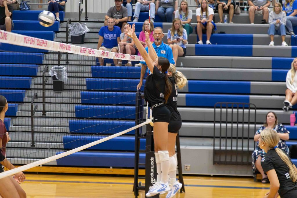 A volleyball player jumps to block a ball at the net during a match, with spectators seated in the background.