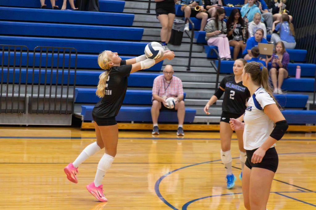 A female volleyball player sets the ball with an upward motion while two teammates look on, in a gymnasium filled with spectators.