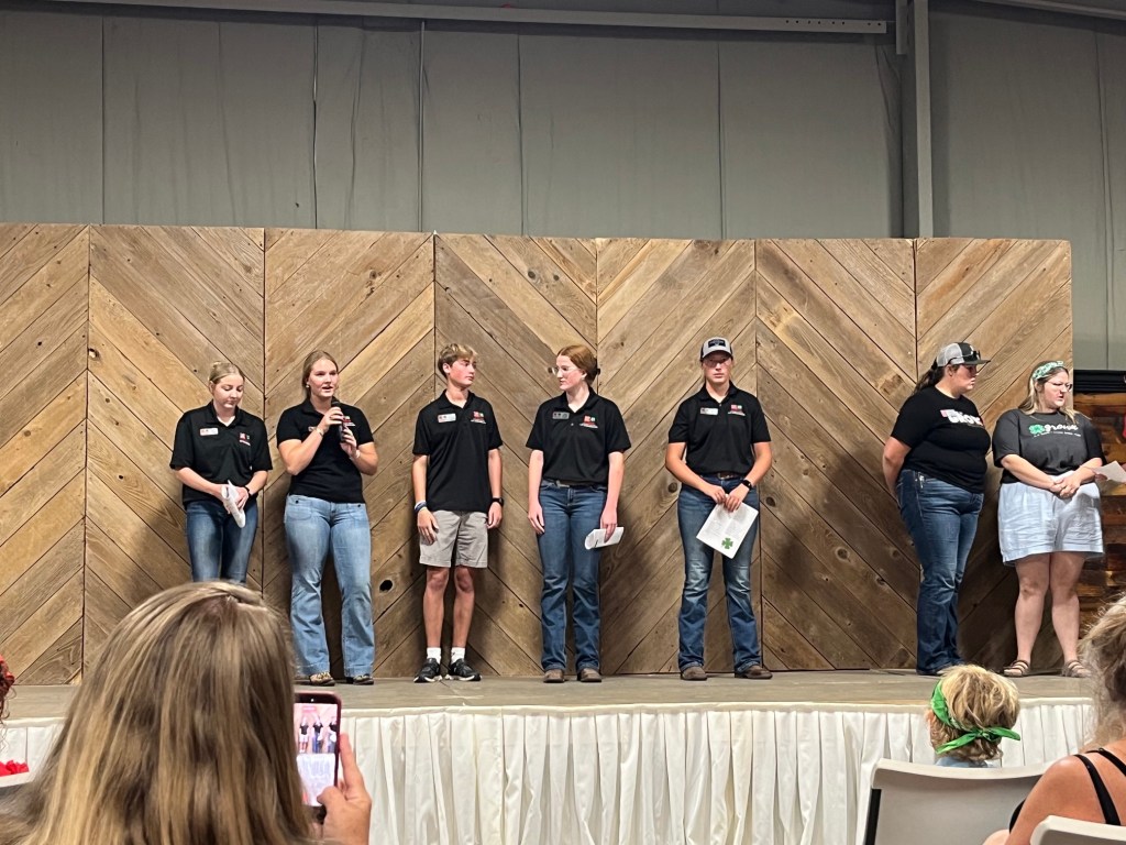 A group of six individuals standing on a stage in front of a wooden backdrop, with one person speaking into a microphone while others listen. They are wearing matching black shirts and appear to be participating in an event or presentation.