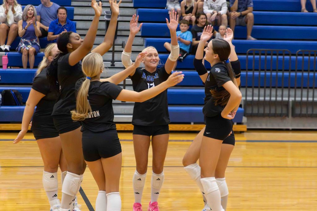 A group of female volleyball players celebrate with raised hands on the court, showing enthusiasm and teamwork, with spectators in the background.