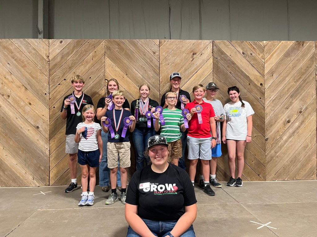 A group of children and teenagers standing on a stage, proudly holding medals and awards, with a wooden backdrop. A girl in front is smiling at the camera, wearing a black t-shirt. The others are arranged in two rows behind her.