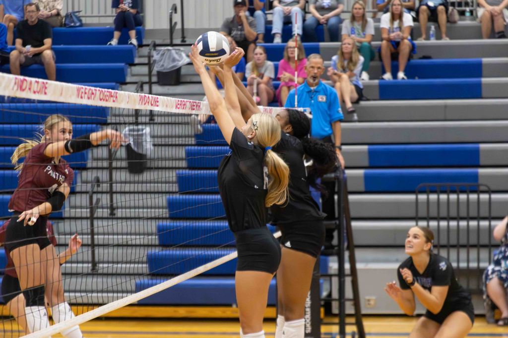 A volleyball match in progress with players from both teams actively engaging at the net. One player is attempting to block a ball while another player prepares to spike. The audience is visible in the background.