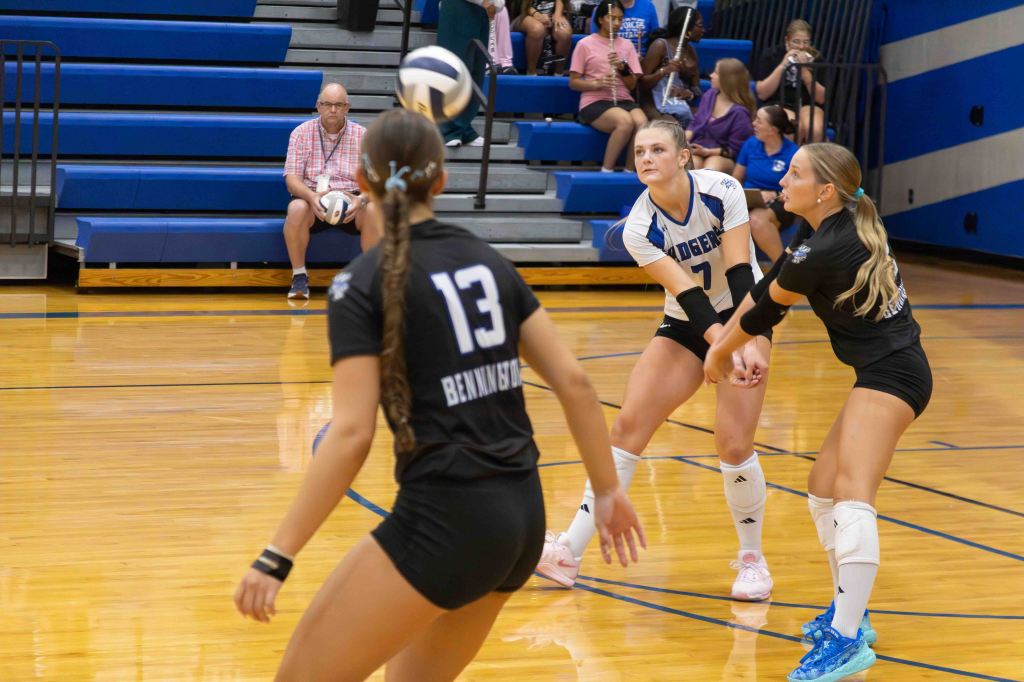A volleyball game in progress, featuring players in black and white uniforms on a gymnasium court. One player is preparing to receive a serve, while another player is poised to pass the ball. A spectator is seated in the background, watching the action.
