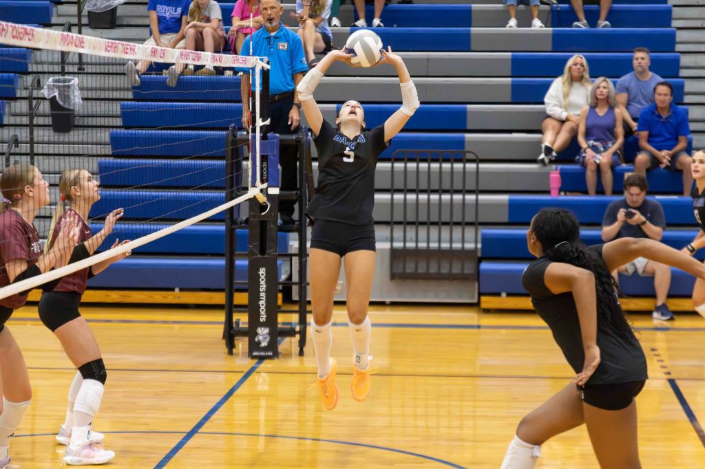 A volleyball player in a black jersey sets the ball for a teammate during a game, with two opponents ready to block at the net and spectators in the background.