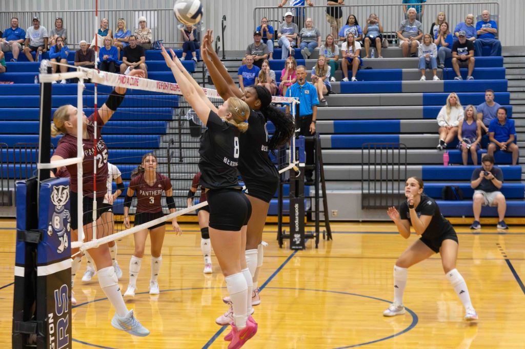A volleyball match in action, featuring players jumping at the net to block a ball. Several spectators are visible in the stands, and players are wearing team jerseys.
