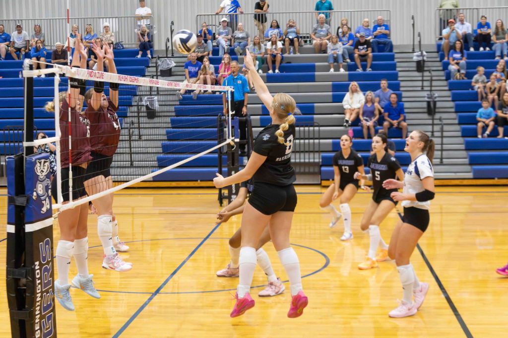 A volleyball game in progress, featuring players in black and maroon uniforms, with a ball being spiked over the net. Spectators are visible in the background, watching the match.