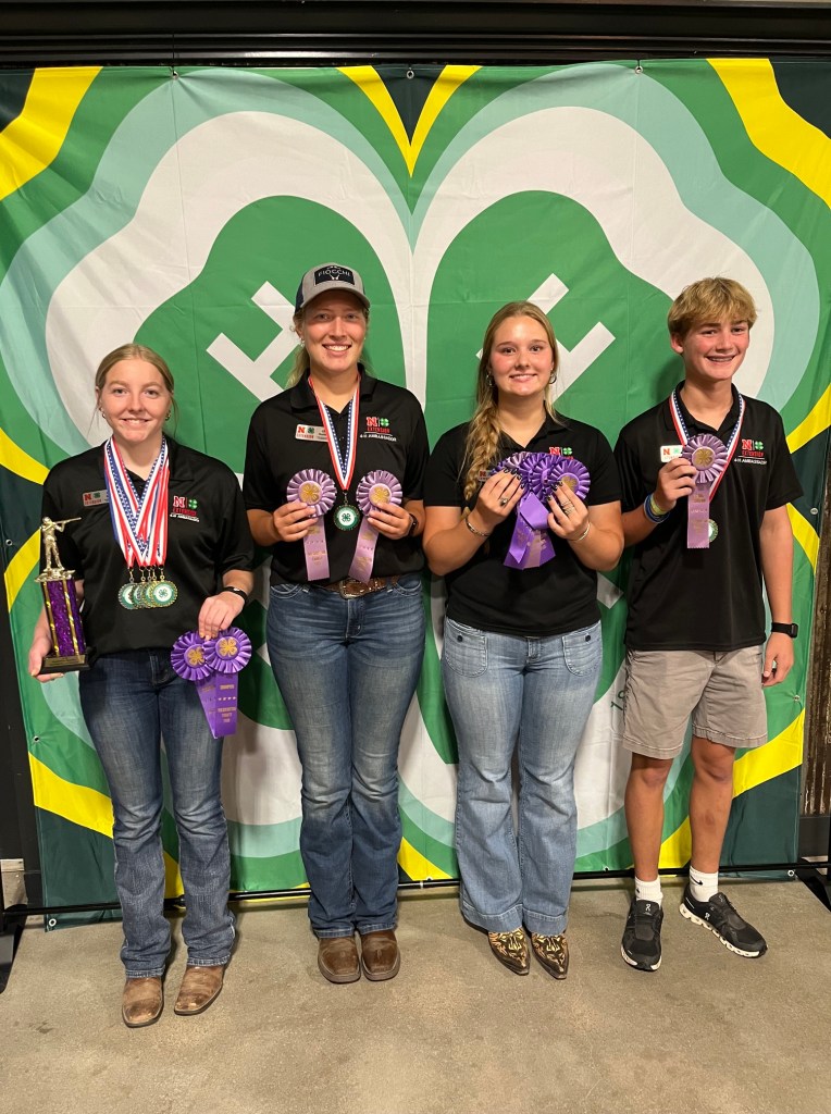 Four young individuals holding awards and ribbons, standing in front of a green and yellow patterned backdrop.