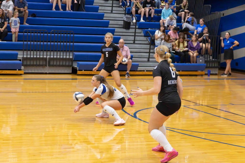 A volleyball player dives to receive the ball during a match, with two teammates nearby. The setting features bleachers filled with spectators.