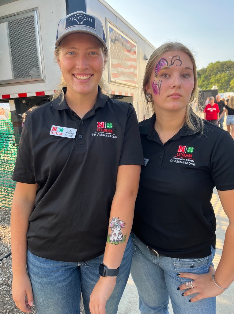 Two young women in black 4-H ambassador shirts posing together outdoors. One has a face painted with a colorful butterfly design, while the other shows a tattoo of a rabbit on her arm.