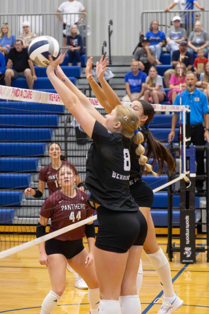 Volleyball players jumping to block a spike at a volleyball match, with spectators in the background.