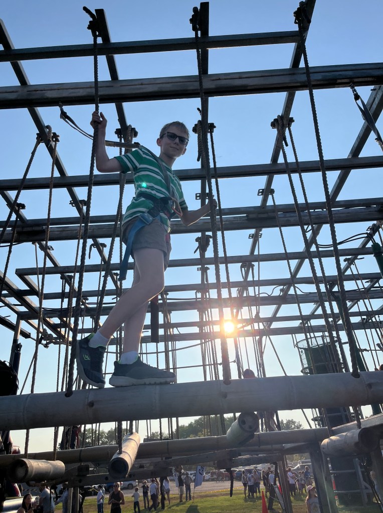 A child wearing sunglasses climbs on a structure made of beams and ropes, with the sun setting in the background and spectators below.