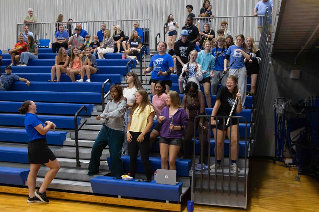 A group of students standing on bleachers in a gymnasium, some holding musical instruments, engaging in a lively activity with a leader in a blue shirt.