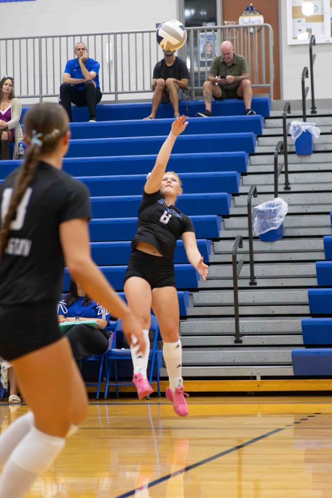 A volleyball player in a black uniform prepares to set the ball in a gymnasium, with spectators seated in the background.
