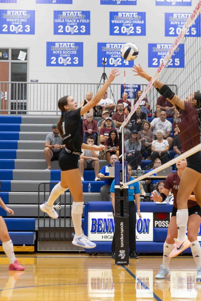 A volleyball player jumping to block a spike at a high school volleyball match, with spectators in the background.
