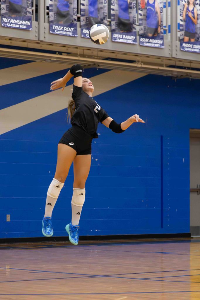 A volleyball player jumping to serve the ball in a gym, wearing a black jersey and white knee pads, with blue walls and banners in the background.