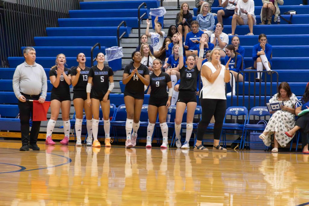 A group of volleyball players enthusiastically cheering from the sidelines during a match, wearing black uniforms with white accents. A coach or official stands nearby with a red clipboard, while spectators watch from the bleachers in the background.