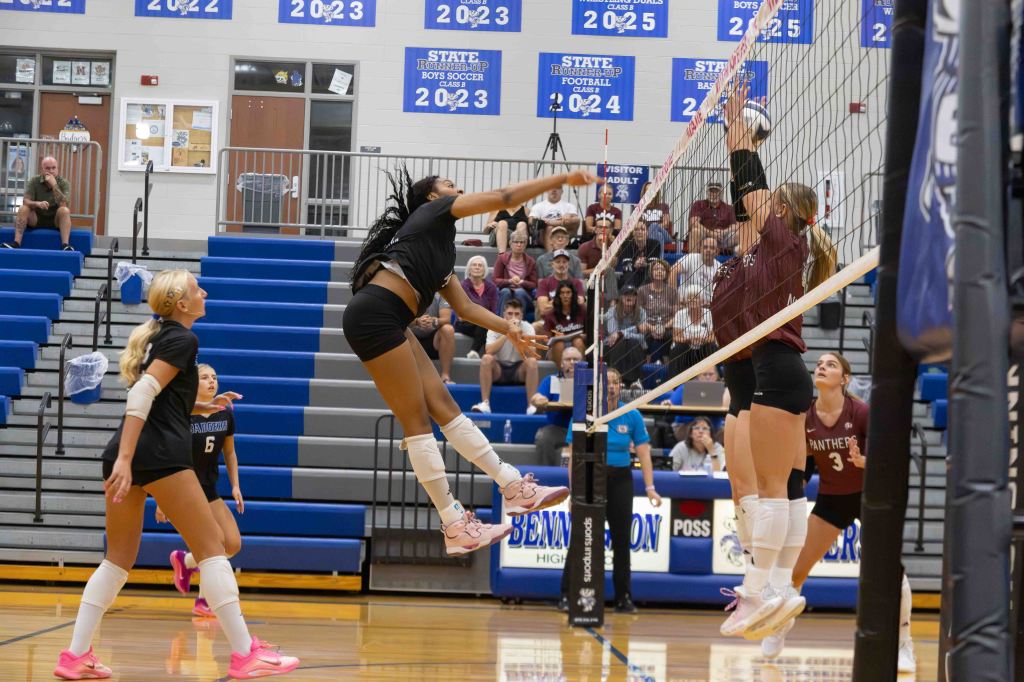 A high school volleyball match in action, featuring players jumping for a spike at the net. Spectators are seated in the background, with a scoreboard visible in the gym.