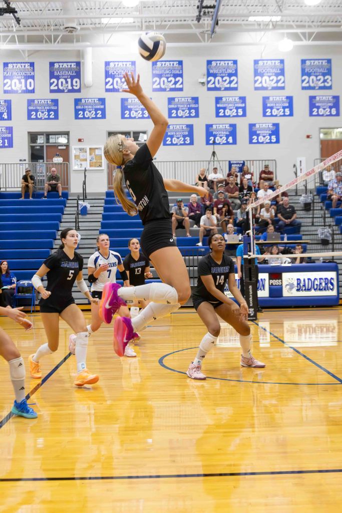 A female volleyball player jumps to set the ball during a match, with teammates positioned around her on the court. The gym features blue bleachers and banners showcasing athletic achievements.