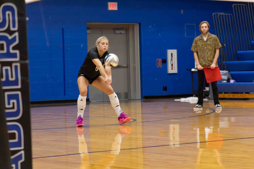 A female volleyball player in a black jersey is preparing to pass a volleyball during a practice session, while a male coach stands nearby holding a clipboard. The gym has wooden flooring and blue walls.