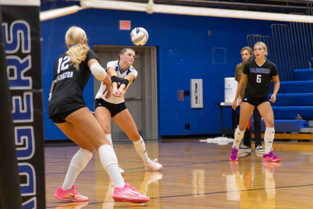 A volleyball match in progress with three players on the court. A player in a black jersey prepares to receive a serve, while another in a white jersey is poised for action. The third player is watching from behind.