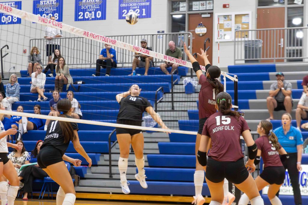 A volleyball game in progress, showcasing players from two teams jumping near the net, with a ball in the air. Spectators are seated in the background, and a banner reading 'WE FIGHT TOGETHER' is visible above the net.