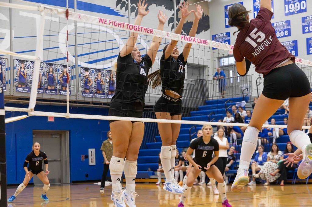 A volleyball match in action, featuring players jumping at the net to block a spike. Two players in black uniforms are attempting to block a player in maroon, while other teammates are positioned on the court watching. The gym is filled with spectators.