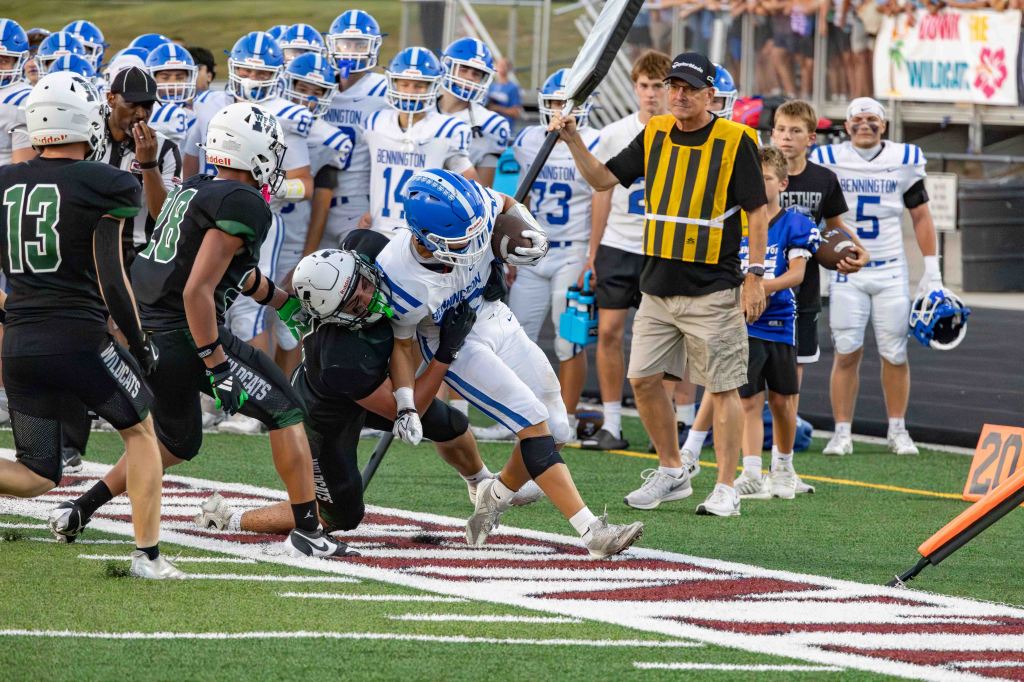 A high school football game scene, featuring an intense play where a player in a blue and white uniform is tackled by a player in a black uniform. Coaches, players, and spectators can be seen in the background, with a scoreboard and yard marker visible.