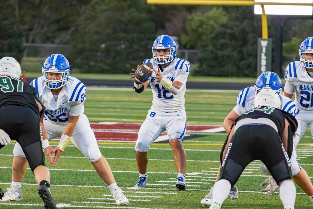 A football quarterback in a white uniform with blue stripes prepares to pass the ball during a game, with teammates and opposing players in action on the field.