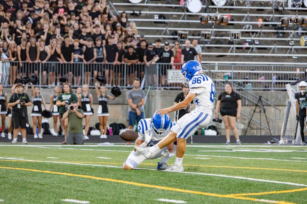 A football kicker preparing to kick a football while a holder supports it, with a cheering crowd in the background at a sports event.