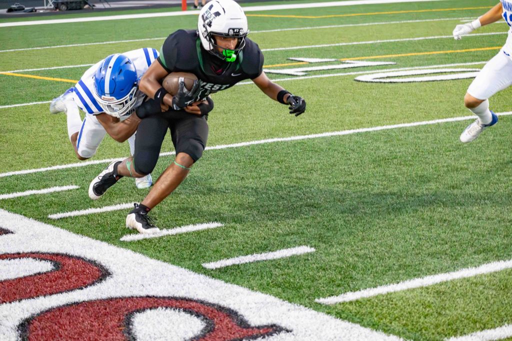A football player in a black uniform is being tackled from behind by an opposing player in a blue and white uniform on a green field.