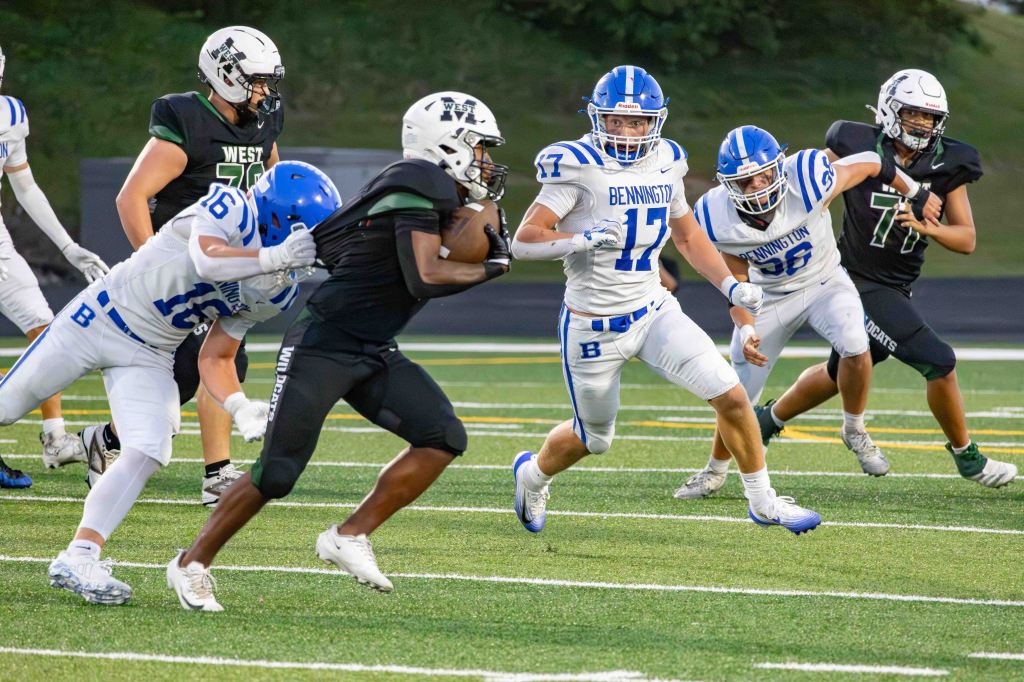High school football game featuring players in blue and white jerseys from Bennington pursuing a player in a black jersey, surrounded by teamwork and dynamic action on the field.