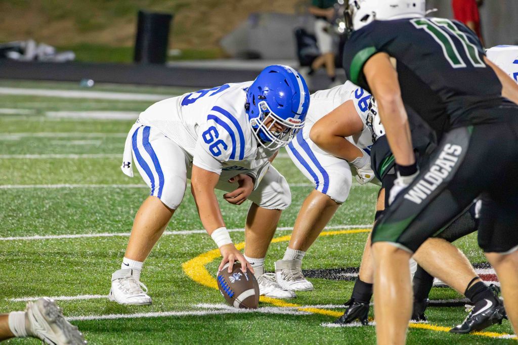 A football player in a white jersey with blue stripes prepares to snap the ball at the line of scrimmage, while opposing players in black jerseys observe the play.