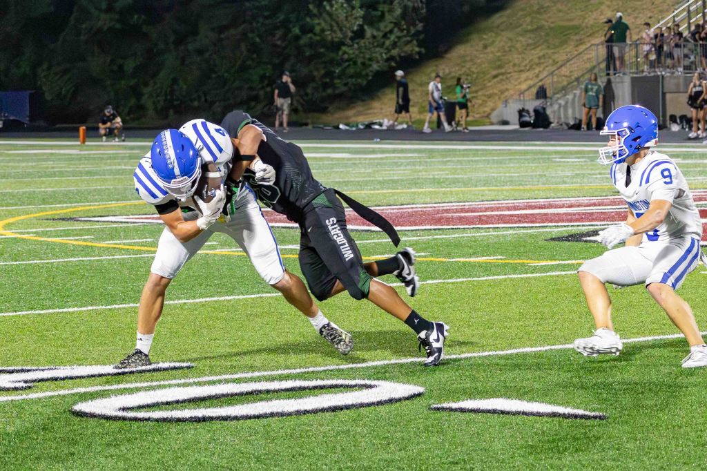 Two football players engage in a tackle on a field, while a third player approaches in the background.