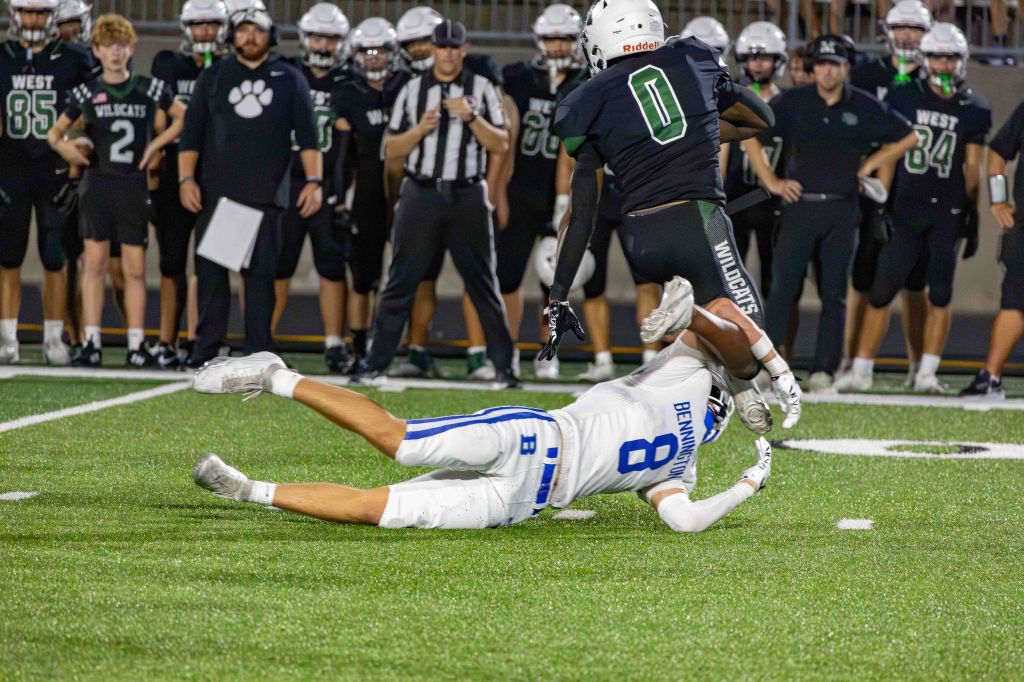 A football player in a white jersey with the number 8 is tackling another player in a black jersey with the number 0 on a green turf field, with several teammates and coaches in the background.