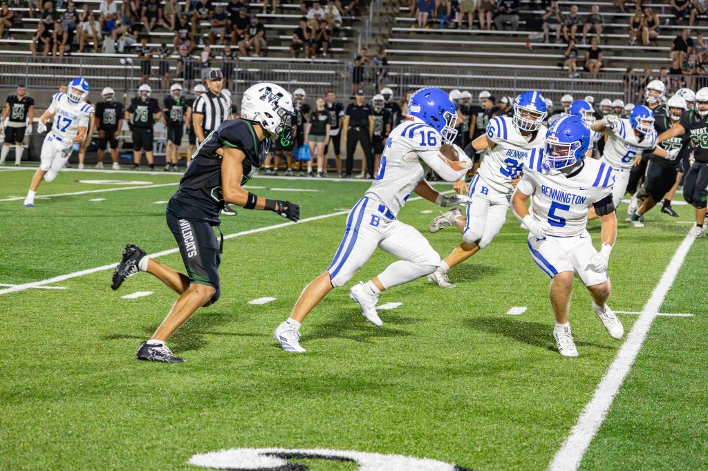 A football game scene with players in white and blue uniforms from Bennington and black uniforms from a different team. One player is running with the ball while being pursued by a defender.