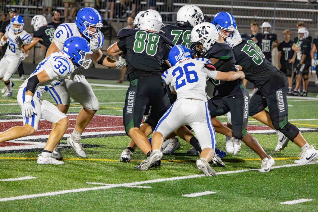 A competitive high school football game featuring players in blue and white uniforms tackling a player in a black uniform on a field.