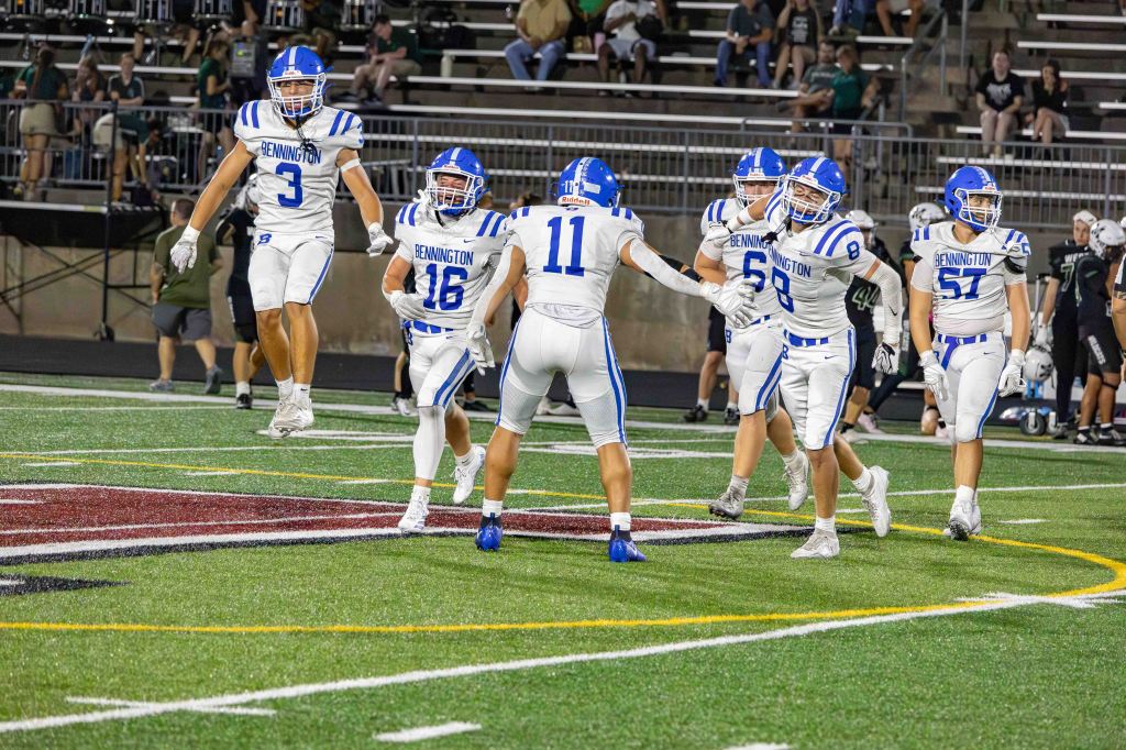 A group of high school football players in white jerseys with blue accents celebrating on the field after a play, with spectators in the background.