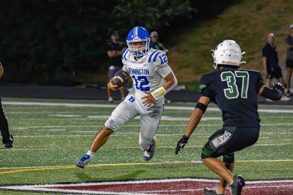 A football player in a white jersey with blue stripes, numbered 12, is running with the ball on a field while being chased by a defender in a black jersey, numbered 37.