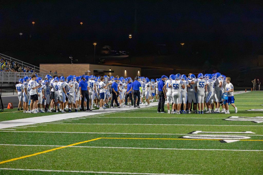 A high school football team gathered on the field at night, wearing white and blue uniforms. Coaches are giving instructions while players huddle together.
