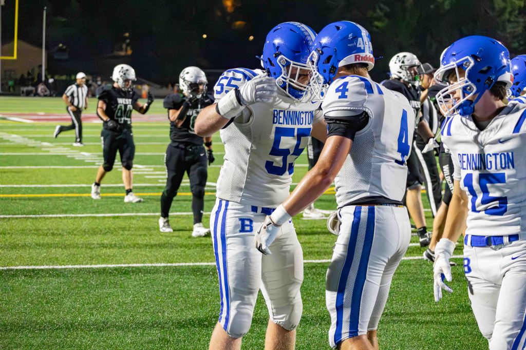 Two football players in white and blue uniforms celebrating on the field, with players in black uniforms in the background.
