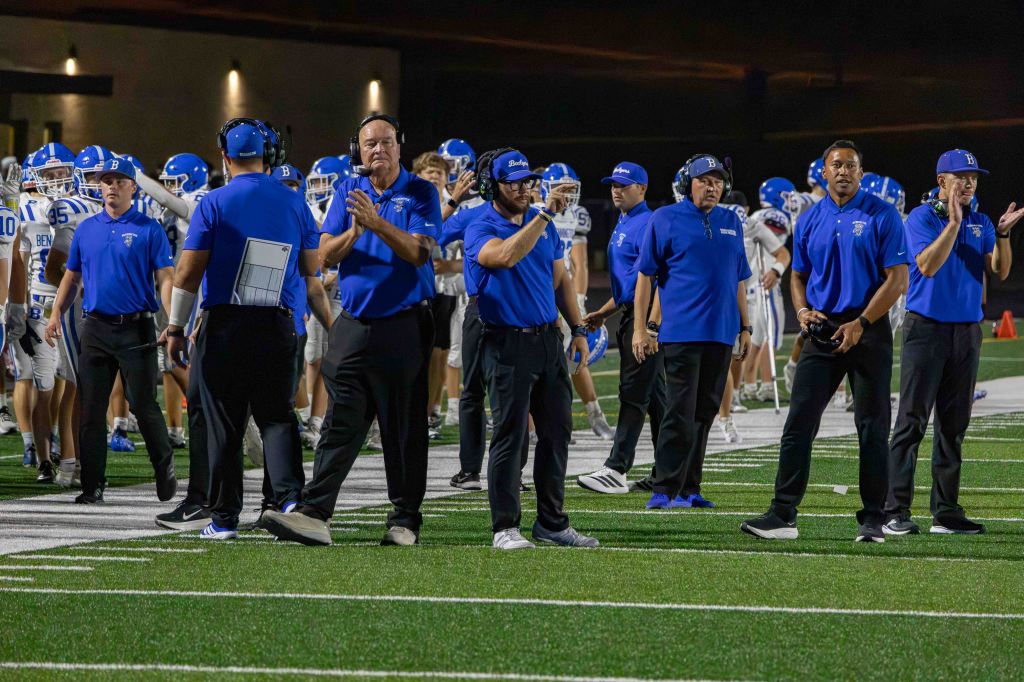 Coaches in blue shirts standing on the sidelines during a football game, observing the players on the field.