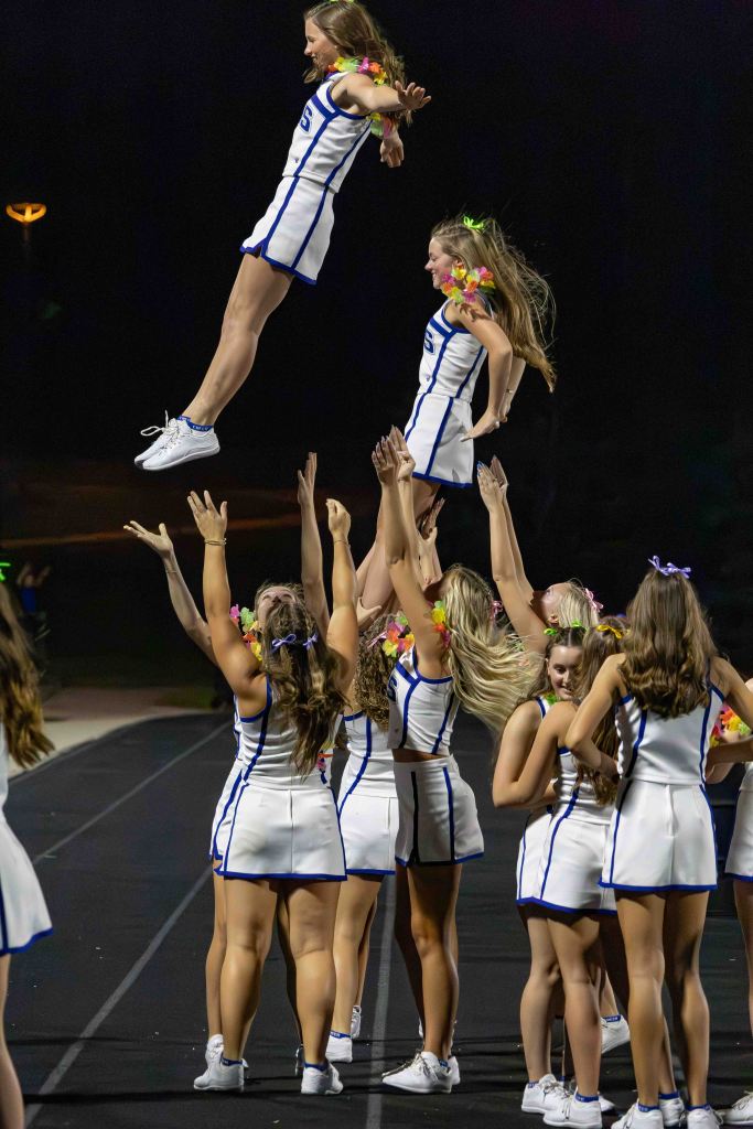 A cheerleader is being lifted into the air by her teammates, who are positioned below her in a choreographed stunt. The background is dark, highlighting the cheerleaders in colorful uniforms adorned with flower decorations.