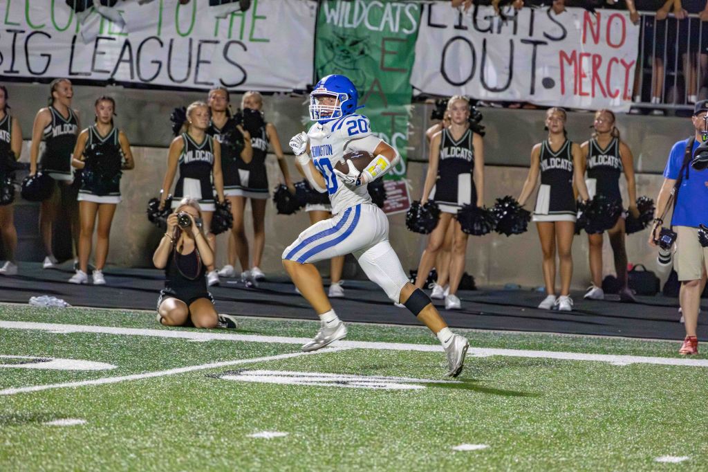 A football player wearing a blue jersey with the number 20 runs on the field during a game, with cheerleaders and banners in the background.